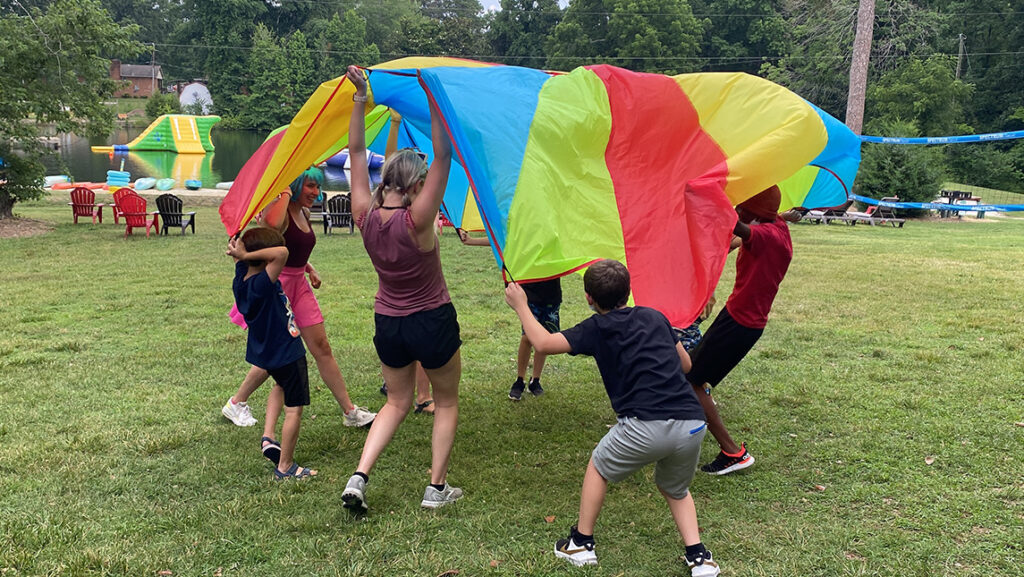 Children and masters students at UNCG play outside with a rainbow-colored parachute